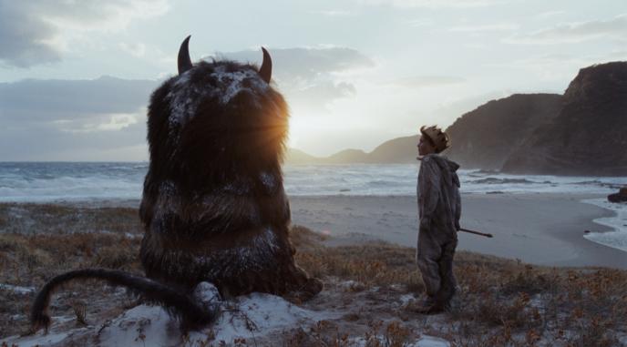 back view of cg animated carol the monster sitting down and max standing next to the monster in front of the sea at a beach at sunset
