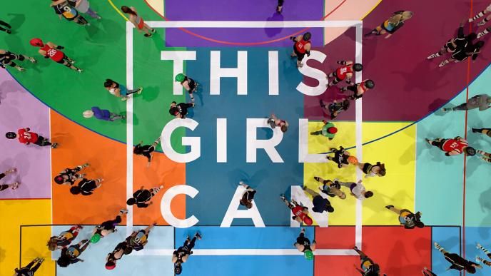 aerial view of a colourful sports court with a large group of girls and women with the text 'this girl can' placed in the centre