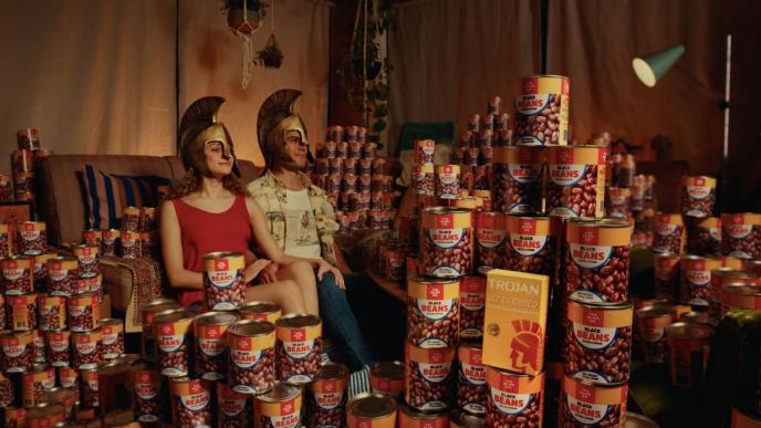 two people sitting in a dimly lit living room wearing roman helmets. the room is full of tinned beans. there is a box of trojan condoms on the right