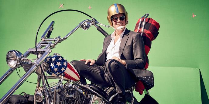 george clooney smiling and looking at the camera as he sits on a harley davidson bike in front of a green screen