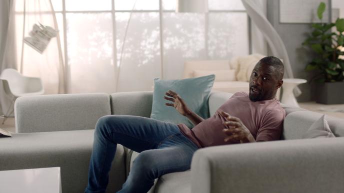 side view of idris elba sitting on a sofa looking into the camera with his hands up