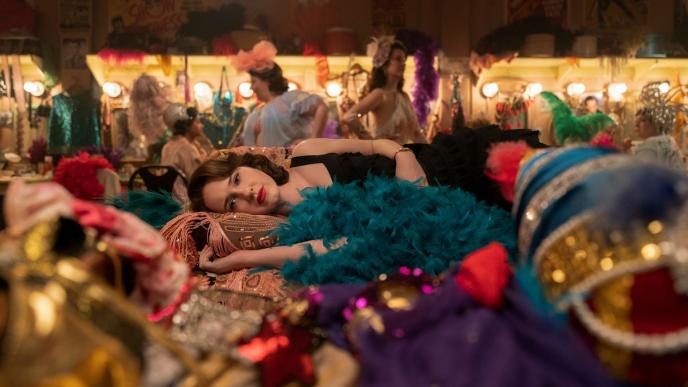 rachel brosnahan as miriam maisel laying on her side on a bed of feather boas and sequins in a dressing room while looking into the camera. there are three show girls standing in the background