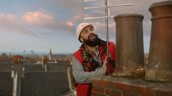 a construction worker standing on top of a roof. he is holding onto a chimney while looking up and smiling