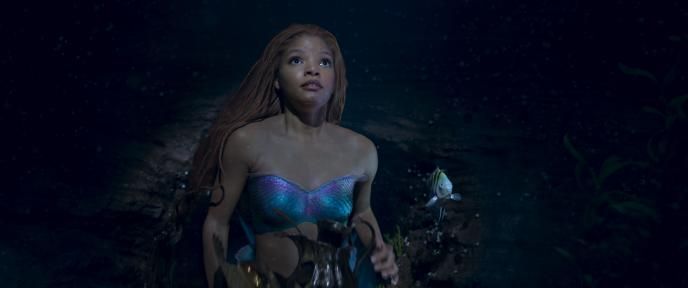 Ariel (Halle Bailey) stops swimming abruptly, looking up, surprised, holding some gold treasures. Flounder floats next to her.
