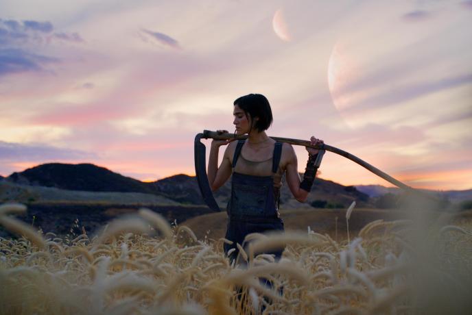 A young woman stands in a field, the sun is setting, she balances a scythe over her shoulders