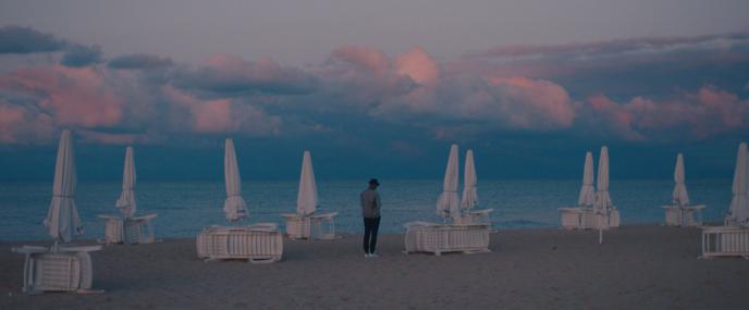a person standing on the beach looking at the sea and mountains that are blankeed with pinky clouds. there are closed umbrellas and tables dotted around the beach