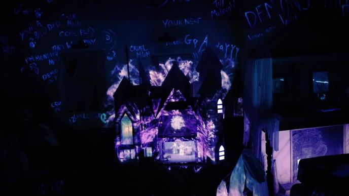 A haunted doll house sitting in a scary room. On the wall beyond the dollhouse are scary words in someone's handwriting
