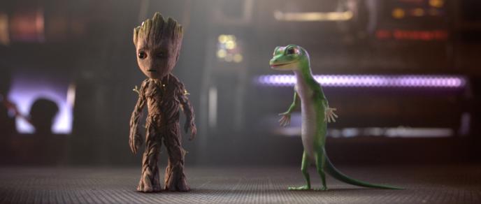 baby groot and geico gecko talking with each other. Geico gecko has its arms open as baby groot is looking away