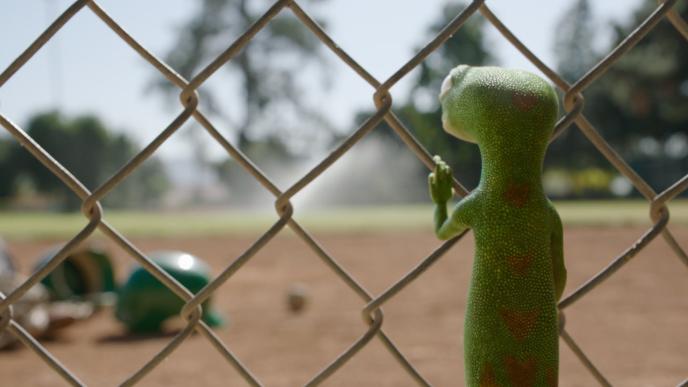 The GEICO Gecko stands at a baseball field, looking through a chainlink fence