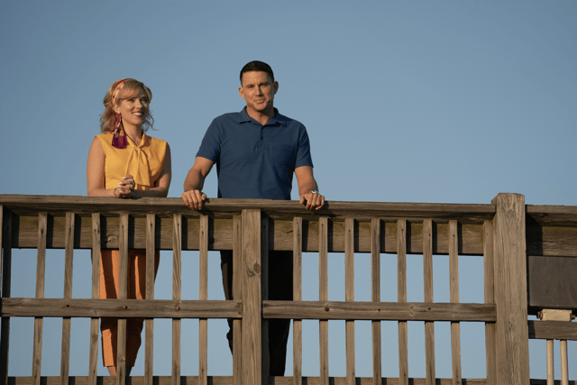 A man and a woman are standing on a wooden platform with a bright blue sky behind them