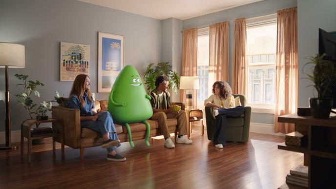 An animated green Cricket Wireless mascot sits on a couch in an apartment with a few people