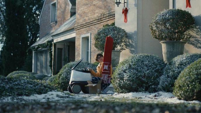 A young boy on his front porch holds gifts a holiday scarf to a delivery robot