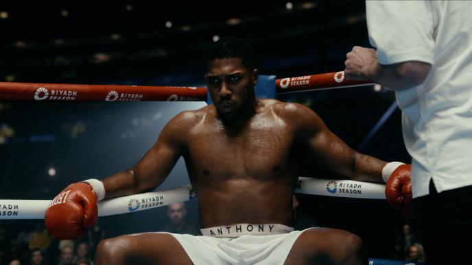 A man sat in a boxing ring with white and red ropes around the edge. 