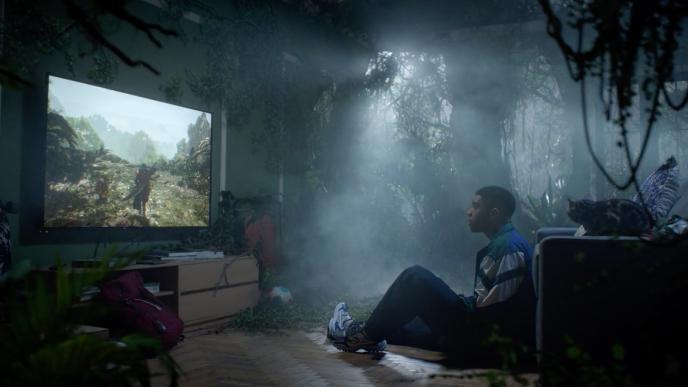 A man plays Monster Hunter Wilds in his living room which is covered in vines