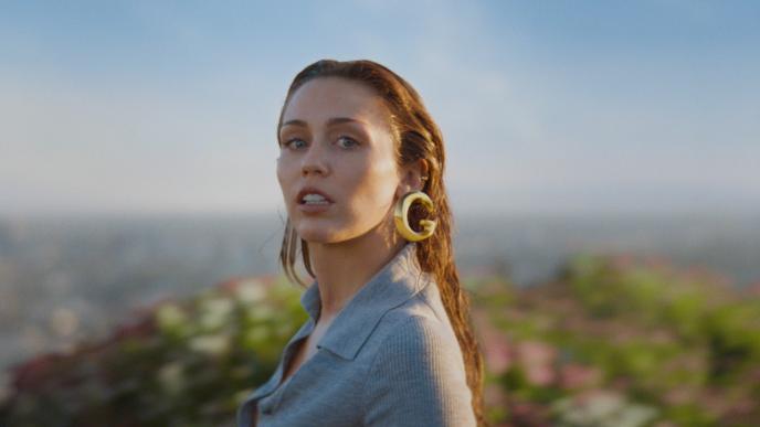 Miley Cyrus stares at the camera while wearing large gold Gucci earring with the LA skyline in the backgroun