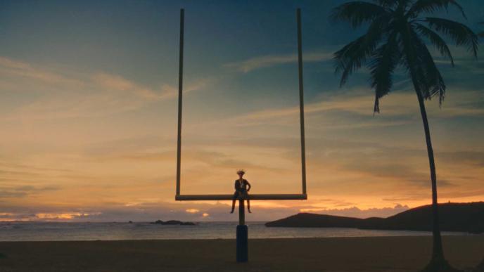 Bad Bunny wearing a straw hat sits on a football goalpost at the beach during sunset, with a palm tree nearby.