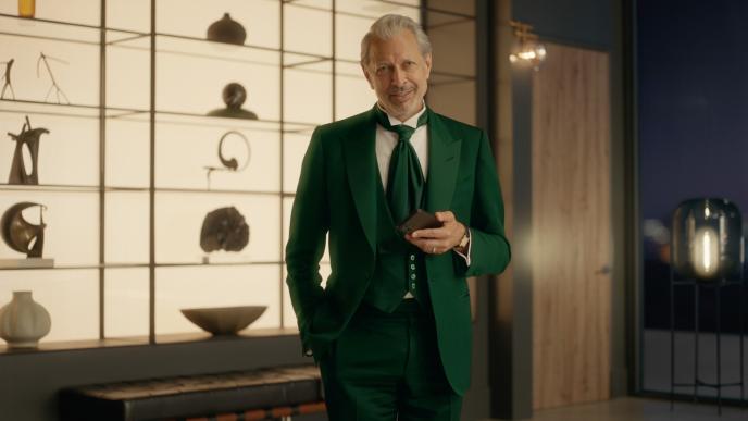 Jeff Goldblum in a green suit stands indoors, holding a phone and smiling before a display of sculptures.