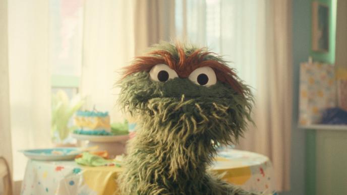 Oscar the Grouch stands in front of a table in a sunny apartment