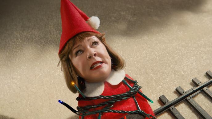 A tiny Melissa McCarthy in an elf-costume figure with a human face lies on snowy ground, tangled in lit Christmas lights beside small toy train tracks.