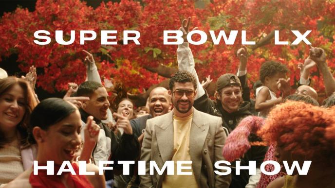 Bad Bunny in sunglasses and a blazer walks through a cheering crowd, arms raised, surrounded by red colored trees behind. The image says "Super Bowl LX Halftime Show"