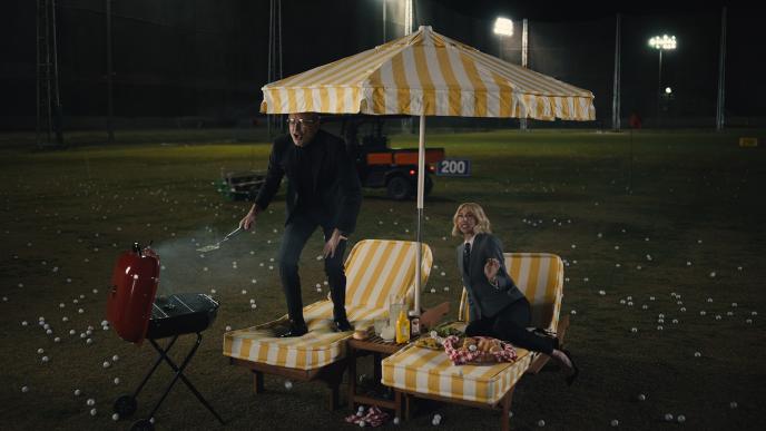 Jeff Goldblum and Heidi Gardner on lounge chairs on a driving range. They are surrounded by golf balls