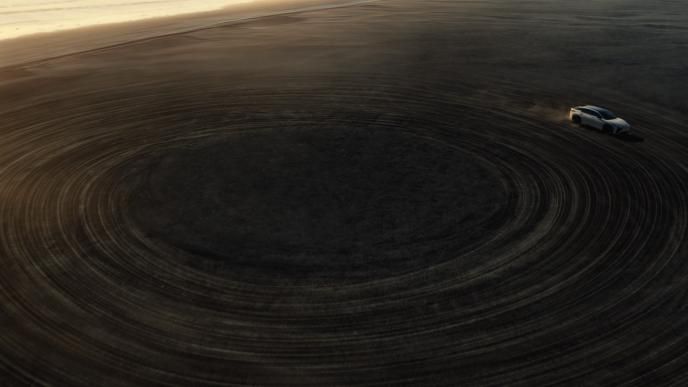 Aerial view of white Lexus SUV driving in large circular tire tracks across dark desert at sunset.