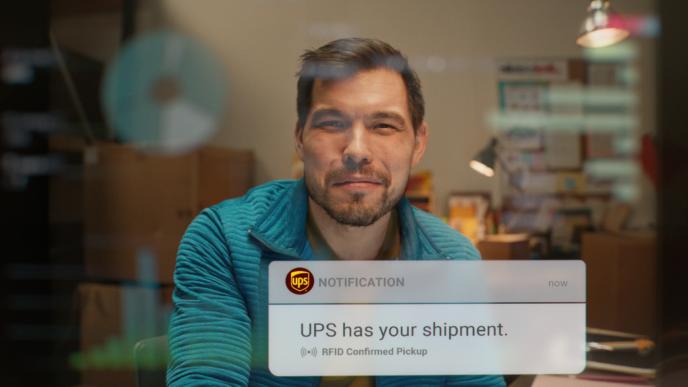 Man looking at camera indoors as a notification reads “UPS has your shipment, RFID confirmed pickup” on screen.