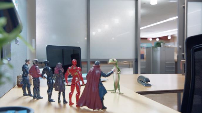 geico gecko mascot talking to six action figures on an office desk