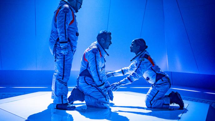 Three astronauts comfort each other, in a blue-lit room