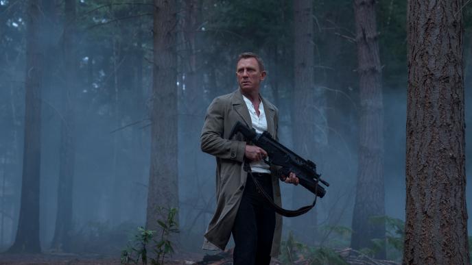 Daniel Craig as James Bond stands in woodland holding a large gun