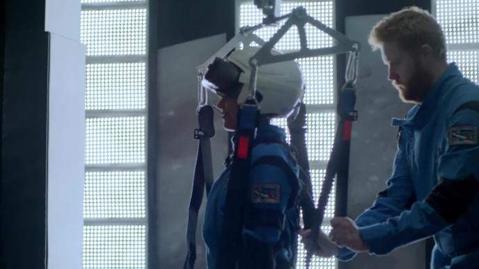 one person helping the other one get into gear with a helmet and braces that is hooked on the ceiling for low gravity effects