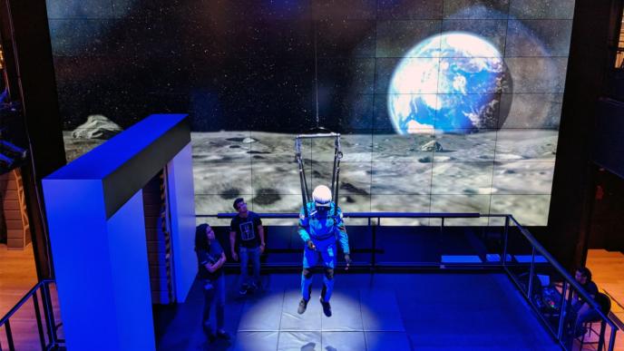 user suspended in air in safety gear whilst supervised by two people with the moon surface and earth in space in the background