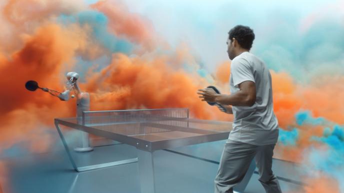 a futuristic table tennis with a person playing against a machine that has a ping pong bat attached to its extended arm. there is colourful mist surrounding the area