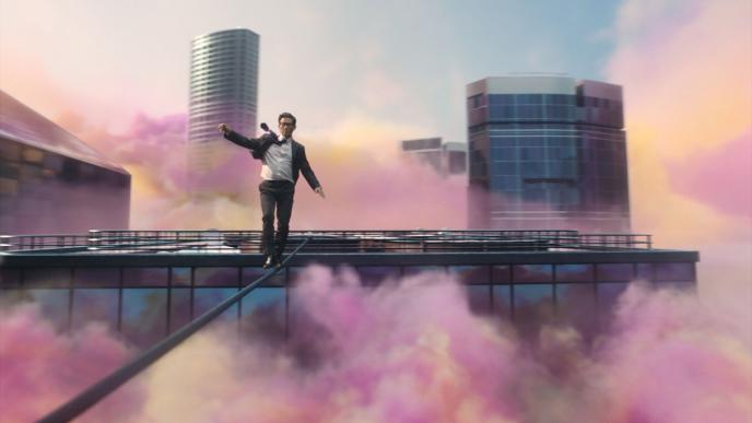 a person in a suit and tie walking on a rope on a rooftop of a cityscape. there is colourful mist surrounding the area