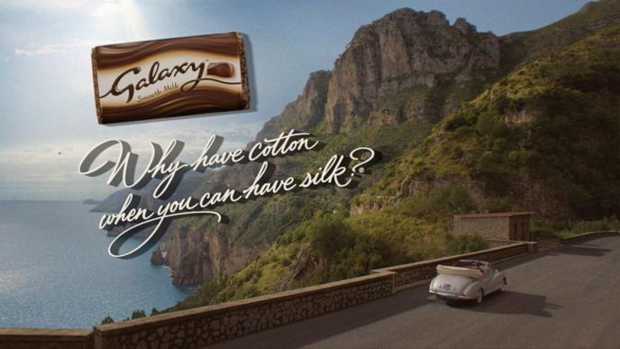a driving by a coast. there is a graphic of a galaxy chocolate bar and text on the upper left corner that says 'why have cotton when you can have silk?'