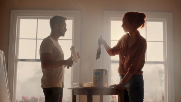 a couple standing in front of windows, above a paint pot preparing to dip their paint brushes 