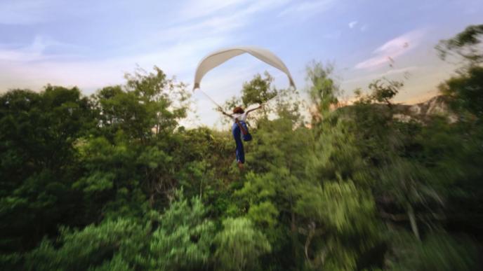 it is dusk. a person is flying down with a parachute amongst forest trees