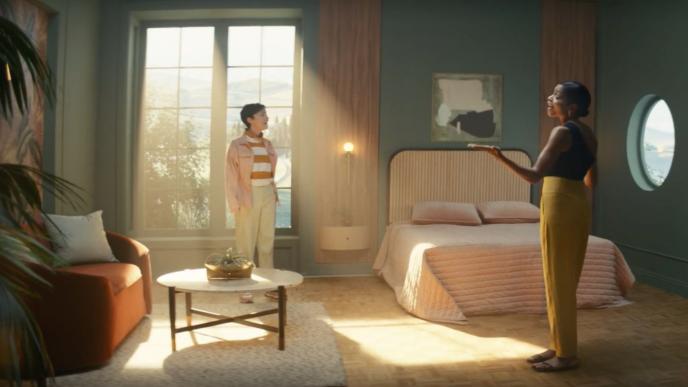 two people standing in a bedroom. one of them is by the bed and the other is in front of the window. there is bright sunbeams coming in through the window