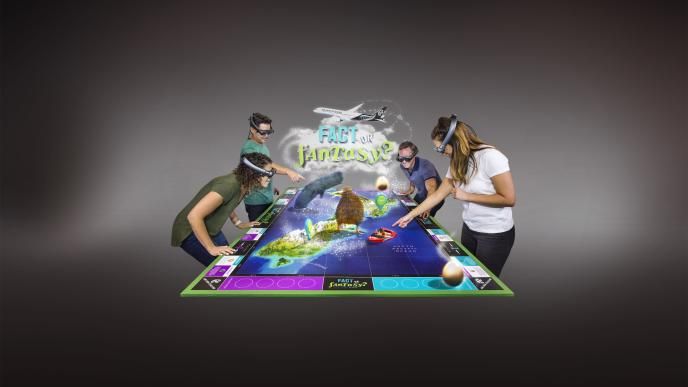 a group of four people in VR headsets standing around and pointing at a virtual board game that has the title ' fact or fantasy' in the centre