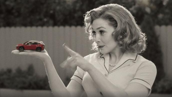 a black and white image of a 50s house wife holding a red car in her palm and pointing at it