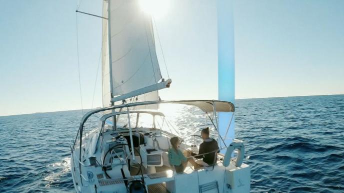 a sail yacht at sea with the sun glimmering ahead. there are two people standing together at the boat's wheel.
