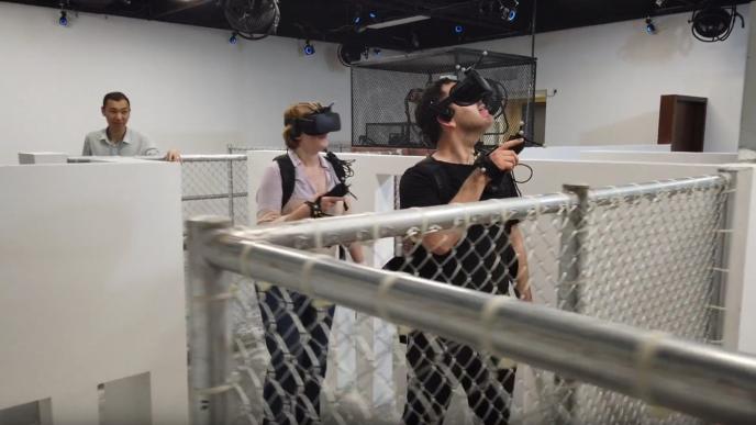 two people wearing vr headsets, holding controllers, walking through an obstacle course while someone supervises them in the background