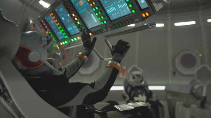 side view of astronauts in a space shuttle. one of them is looking up at a screen