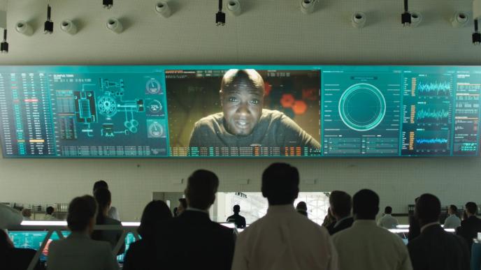 back shot of people in a rocket lab looking at a screen. there is a person on the screen speaking to the conference. there are charts and graphics on each side of the screen