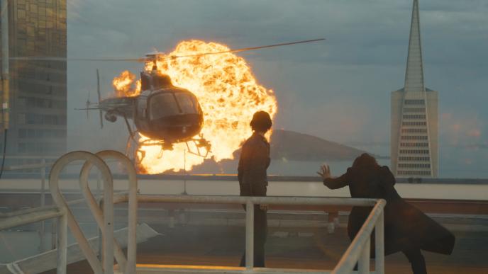 Keanu Reeves and Carrie Ann Moss stand on a rooftop as a helicopter explodes