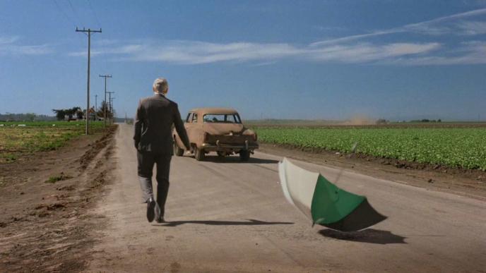 actor george clooney with his back towards the shot. there is an umbrella upturned on the side of the road. a car is driving on