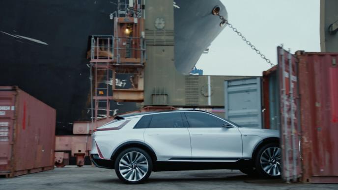a general motors car driving into a shipping container in front of a dock. a bulbous bow and chain anchor is visible in the background