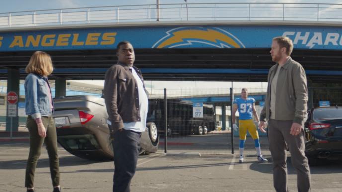 three people standing in front of a flipped car. there is an american football player in the background