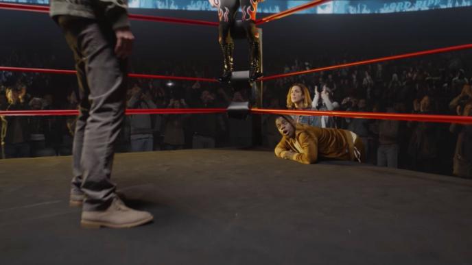 perspective shot from inside of a fight ring. there are two people's legs visible in the ring. the pair of legs visible on the right are firmly stood on the ground. the other pair of legs are hovering mid air. three people are in the corner looking up in awe