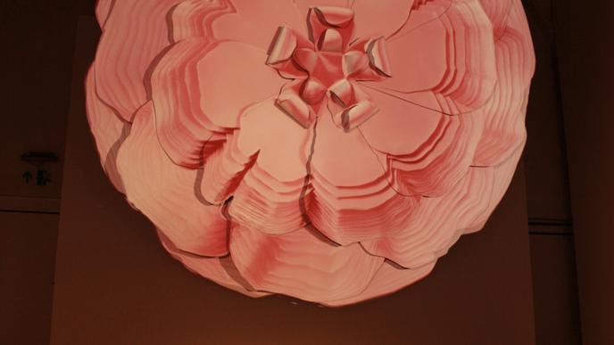 an exhibition of a projection of a graphic origami rose. it is in a dimly lit room and there are people gathered at the bottom of the wall looking down into a projector